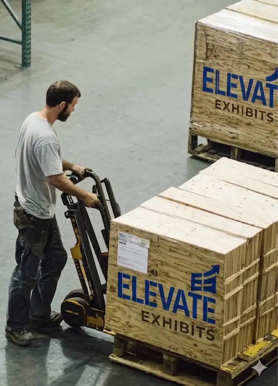 Man operating a pallet jack in a warehouse, moving crates labeled Elevate Exhibits, showcasing trade show logistics and preparation services.